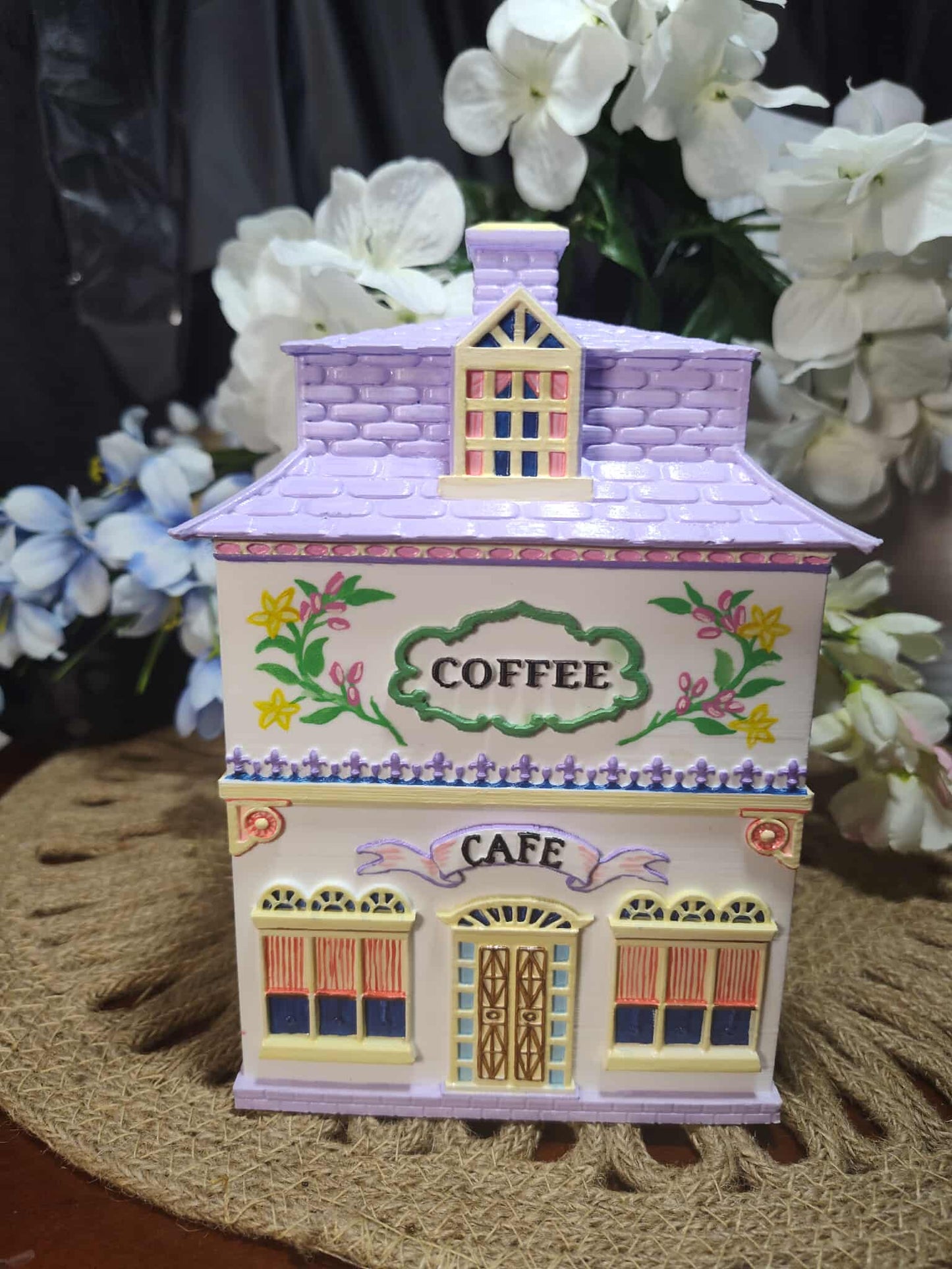 Decorative coffee canister shaped like a small building with flowers and 'Coffee Cafe' text, placed on a textured surface with flowers in the background.