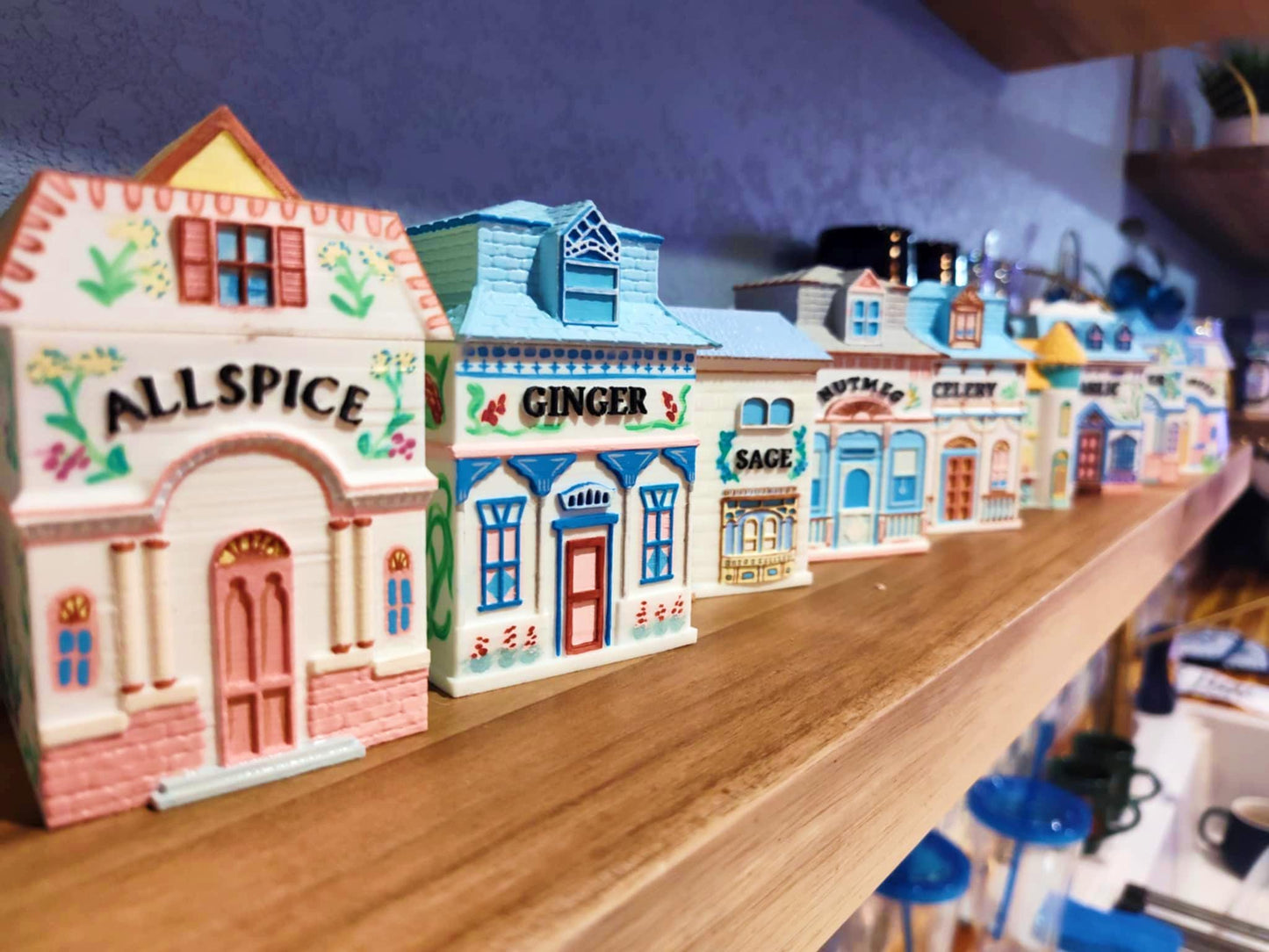 Row of open, 3D printed, hand-painted house-shaped spice jars labeled Allspice, Ginger, Sage, Nutmeg, Celery, and more, inspired by Lenox designs, displayed on a wooden shelf.