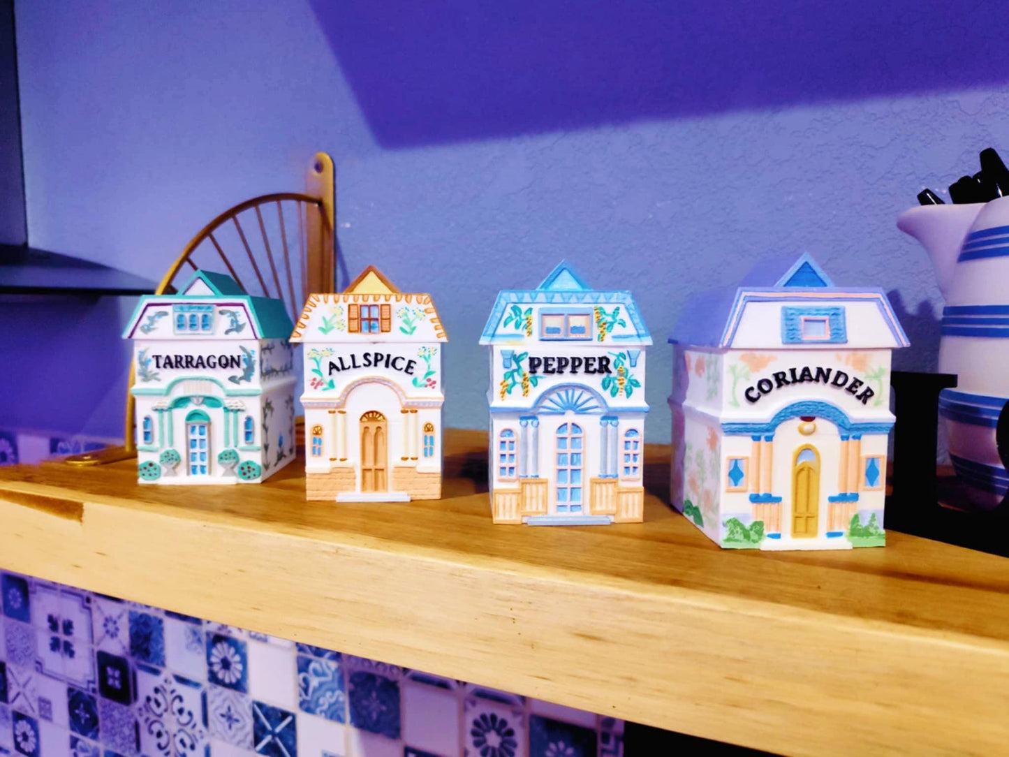 Four 3D printed, hand-painted spice jars shaped like miniature houses, labeled Tarragon, Allspice, Pepper, and Coriander, styled after vintage Lenox village designs and displayed on a wooden shelf.