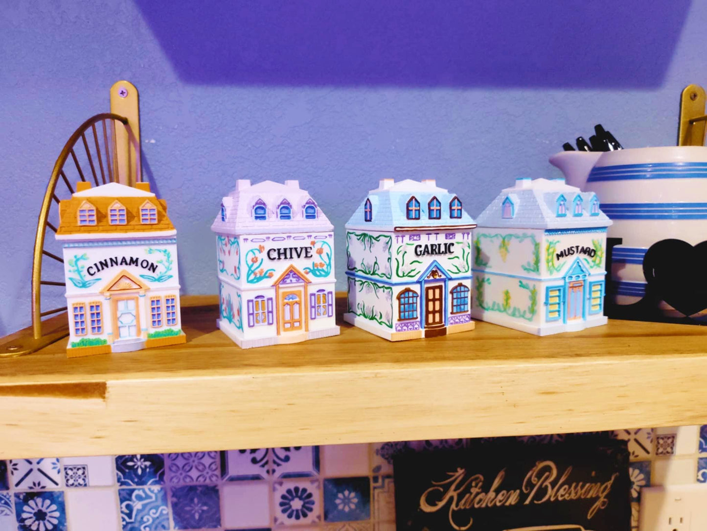 Four 3D printed, hand-painted spice jars shaped like miniature houses, labeled Cinnamon, Chive, Garlic, and Mustard, inspired by vintage Lenox village designs and displayed on a wooden shelf.