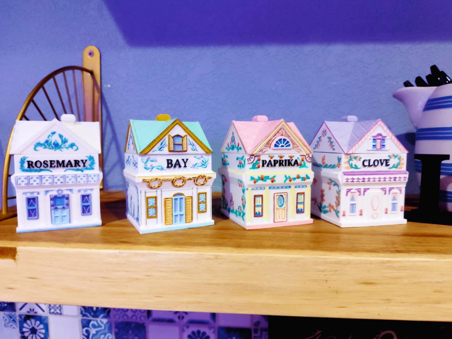 Four 3D printed, hand-painted spice jars shaped like miniature houses, labeled Rosemary, Bay, Paprika, and Clove, inspired by vintage Lenox designs and displayed on a wooden shelf.