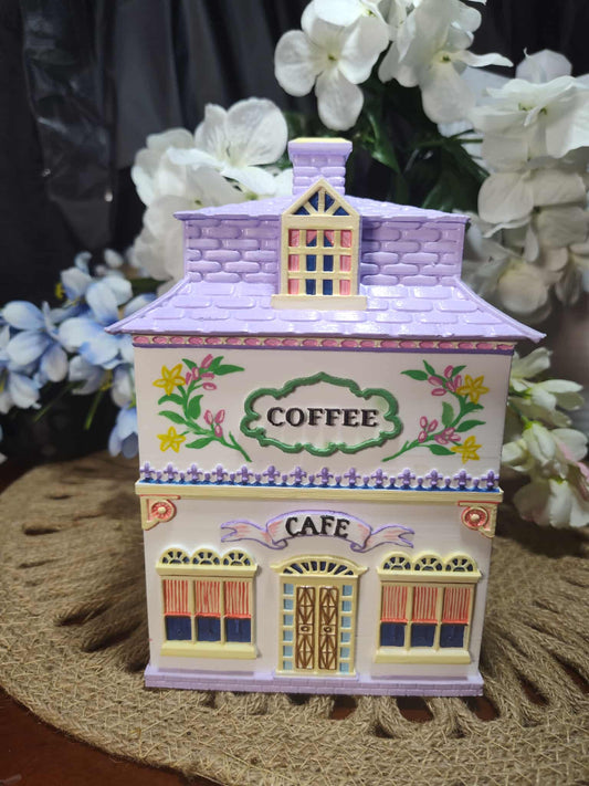 Decorative coffee canister shaped like a small building with flowers and 'Coffee Cafe' text, placed on a textured surface with flowers in the background.